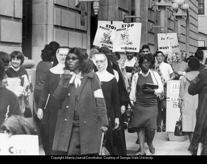 a black-and-white photograph depicting protesters of different ages and ethnicities, including three nuns in traditional habits, demonstrating in front of a building in downtown Atlanta. Some protesters are holding signs that either read STOP POLICE KILLINGS or JIM CLARK MUST GO.